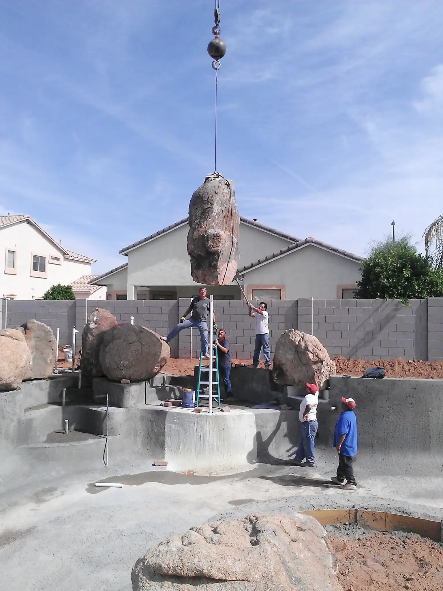 A crane delicately lowers a large boulder into place while a crew constructs a water feature in a new pool build, illustrating the precision and creativity in pool and spa projects.